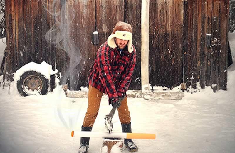 Man wearing a plaid jacket and hat outside in the snow chopping a large pretend cigarette.