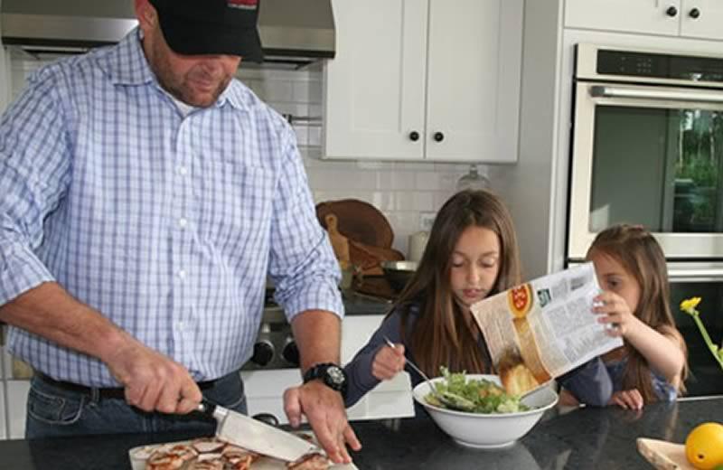 Man wearing hat and plaid shirt cooking with kids in kitchen.