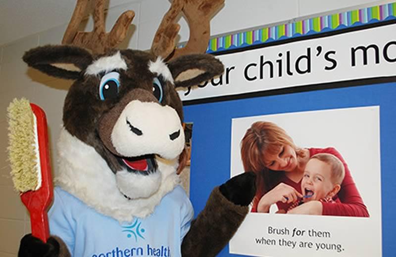 Spirit the caribou mascot holding a giant red toothbrushin front of poster