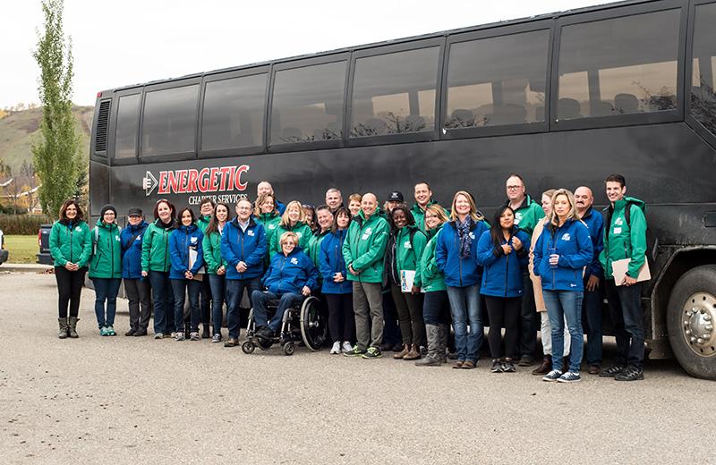 A large group of people in blue or green BC Winter Games jackets stand in front of a bus.