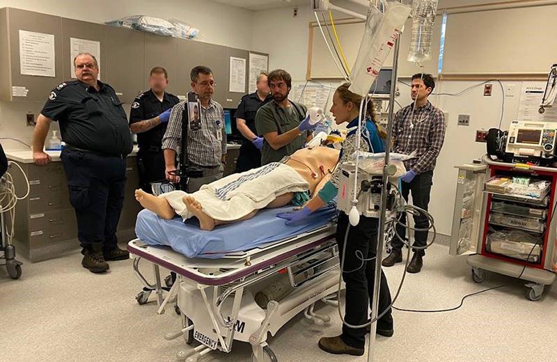 A group of paramedics and health care professionals stand in a medical room around a simulation mannequin.