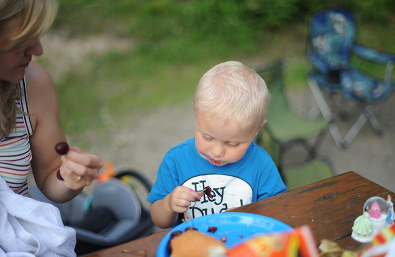 A toddler boy looks at a cherry he is eating at a picnic table while his mom looks at him. 