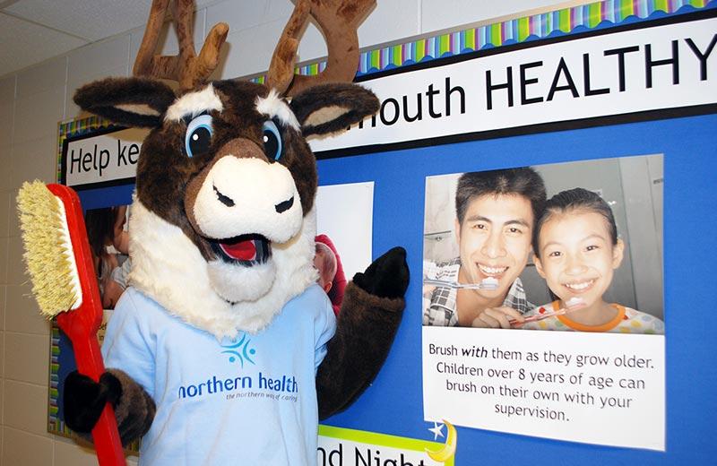Northern Health's caribou mascot holds a giant toothbrush in front of a display promoting mouth health. 