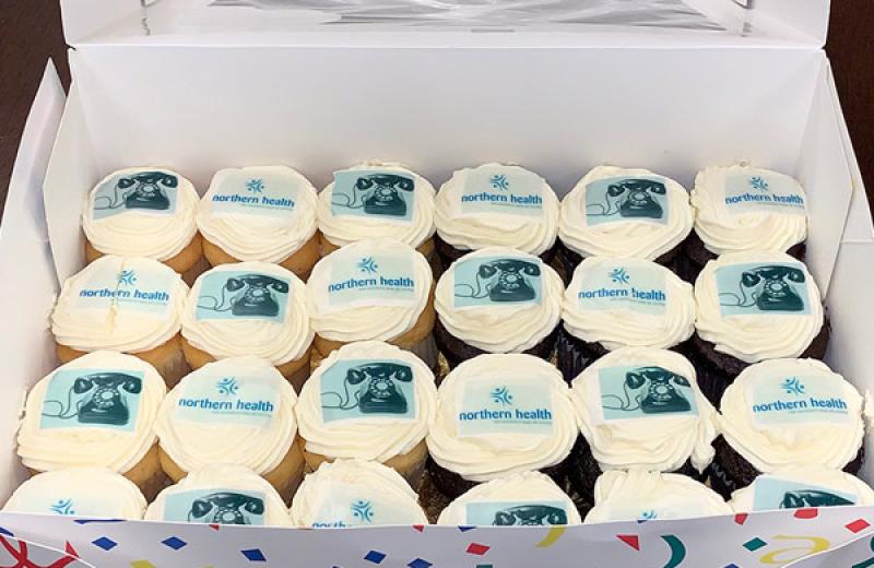 A box of cupcakes that are decorated with the Northern Health logo and pictures of old phones.