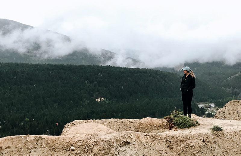 A woman stands at the edge of a cliff, forested mountains touching clouds in the background. 