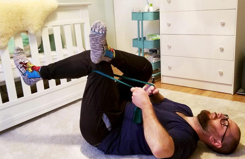 A man lays on the floor,  using a physio band to stretch his hamstrings.