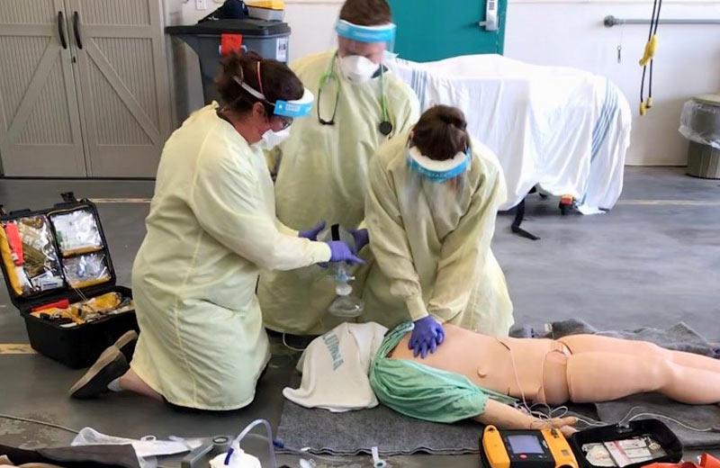 Three people wearing personal protective gowns and masks perform CPR on a simulation dummy. 