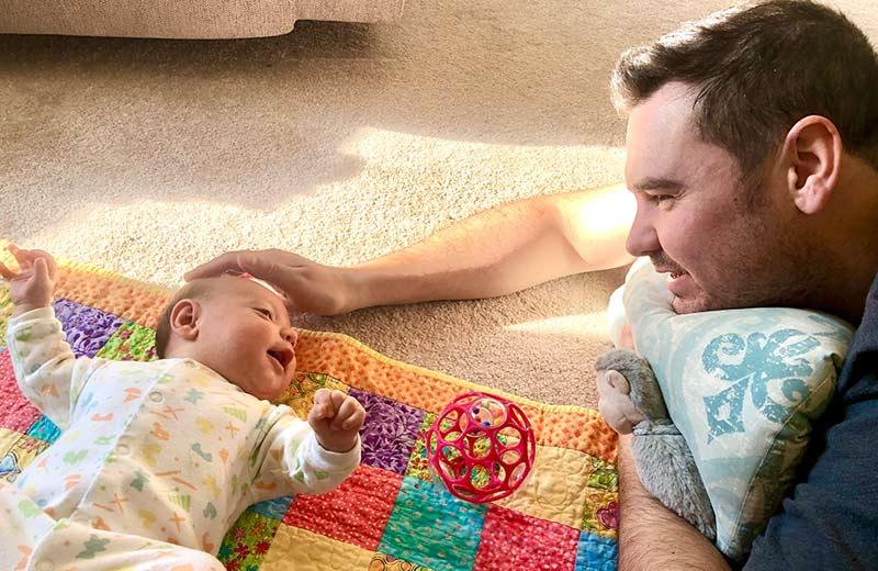 A baby lays on a quilt on the floor, smiling up at its father who looks down and smiles back.