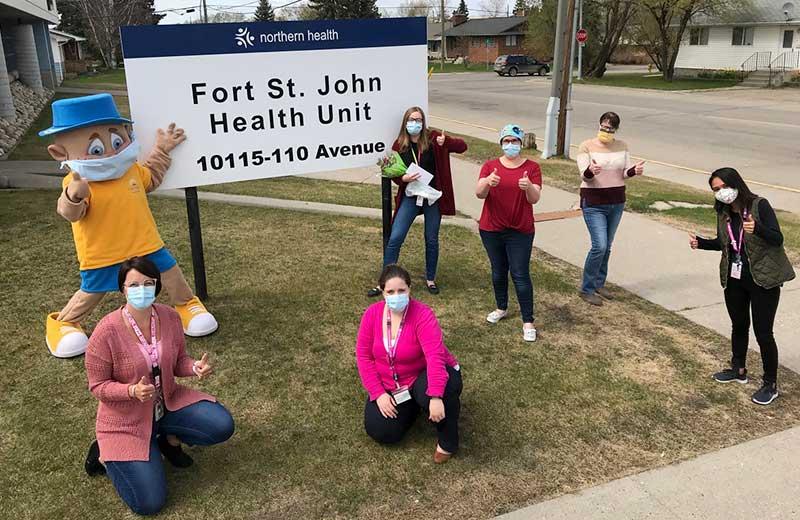 Six nurses and a mascot pose, wearing masks and giving thumbs up signs, outside the Fort St. John Health Unit.