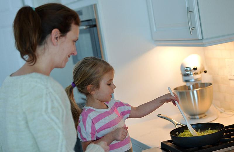 In a kitchen, a woman holds a young girl who stirs food in a pan on the stove.