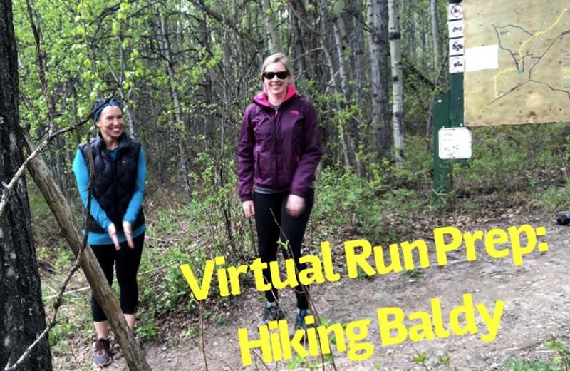 Two women in hiking apparel smile into the camera as they hike a wooded trail.