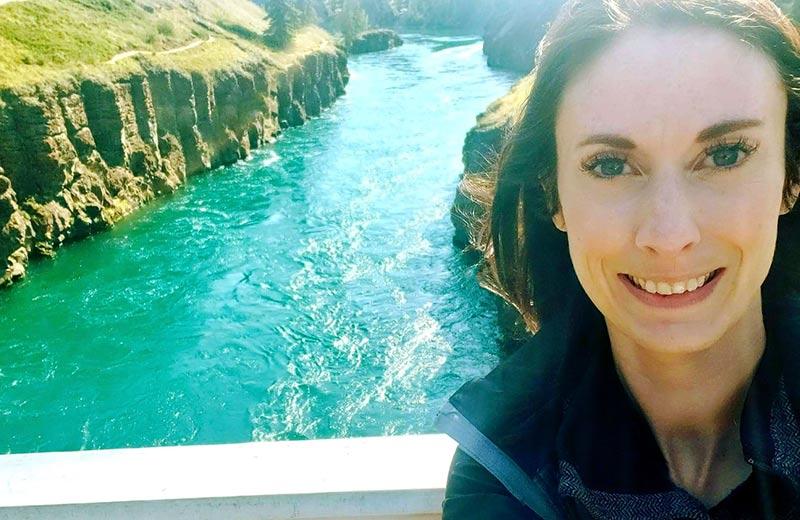 Smiling woman taking a selfie on a bridge with a beautiful teal river behind her.