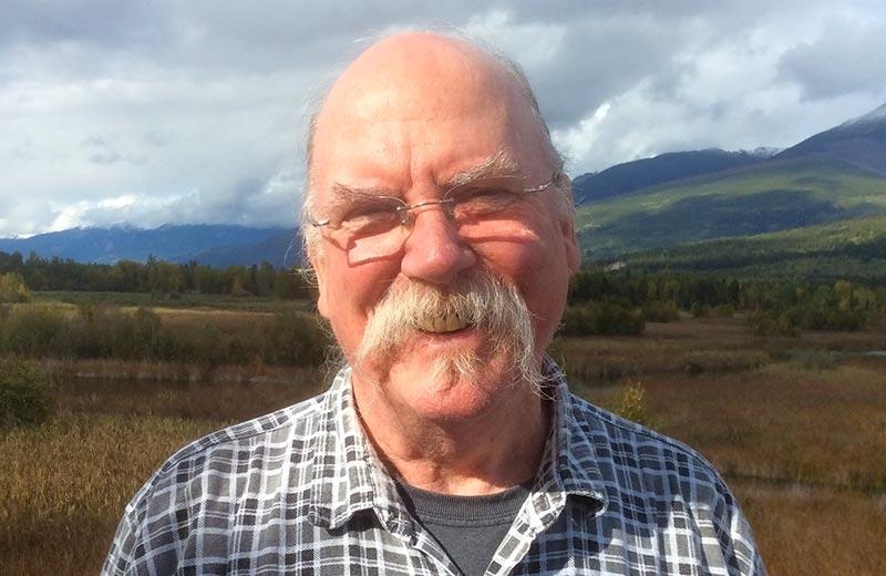 Elderly man with glasses and a large moustache stands in field with mountains in the background
