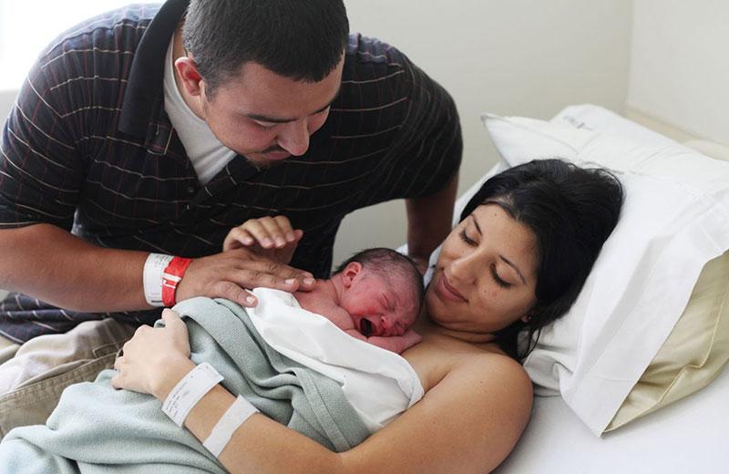 Mother lying in hospital bed holding newborn baby, with male partner watching over.