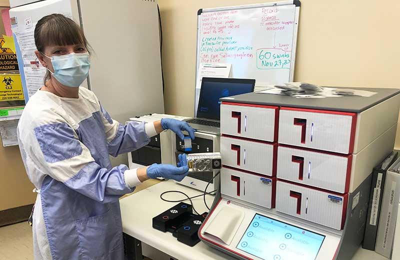 Woman in lab clothes and mask stands next to lab equipment