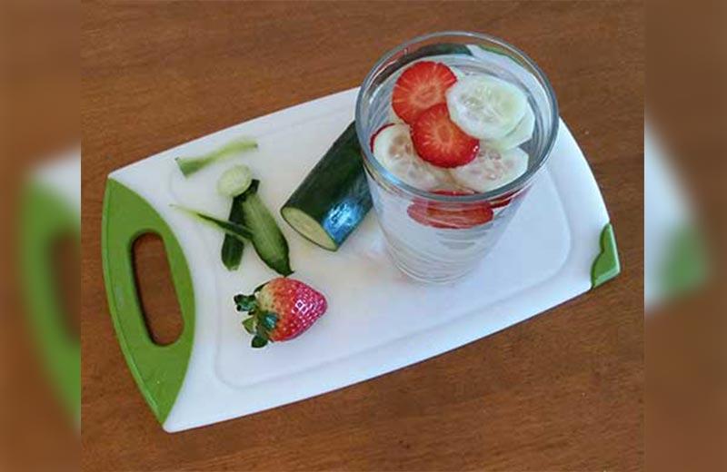 A cucumber and strawberry on a cutting board with slices in a glass of water.