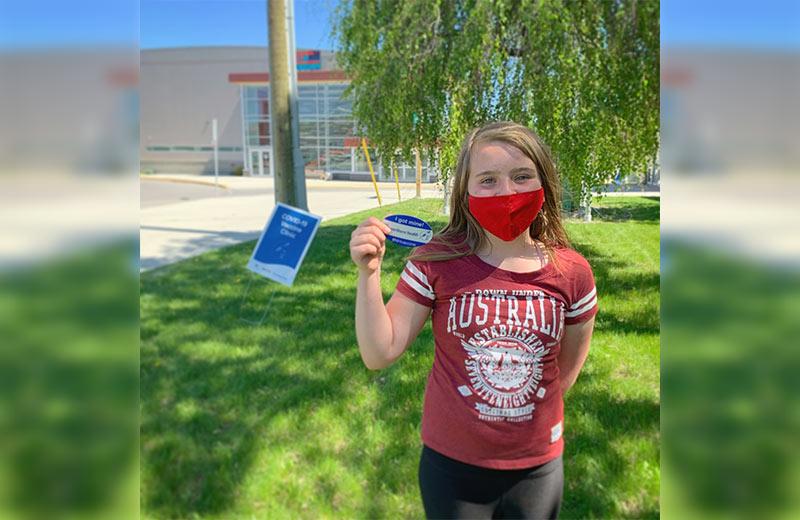 Young girl wearing a red mask while standing on the grass holding her COVID-19 vaccination sticker