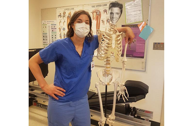 Woman in scrubs stands next to skeleton.