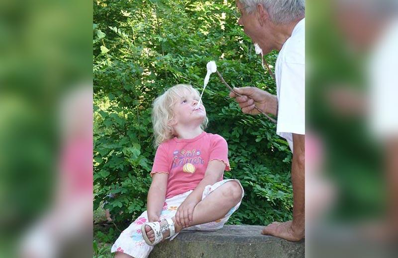 Child sitting on a log eating a marshmallow from her grandfather.