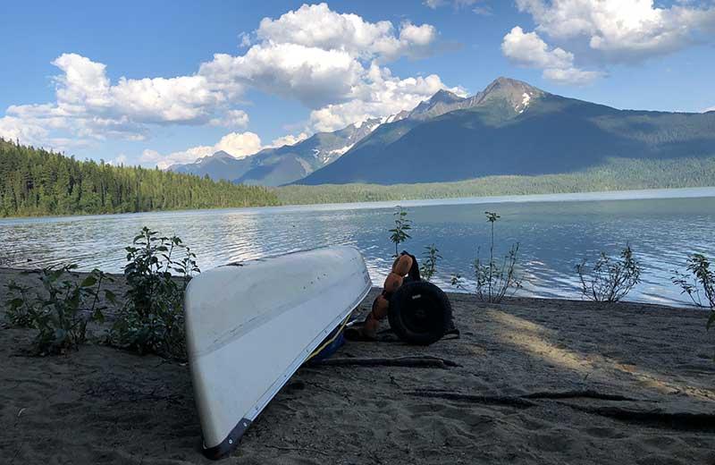 Lake view with a canoe resting on a sandy beach with mountains in the background.