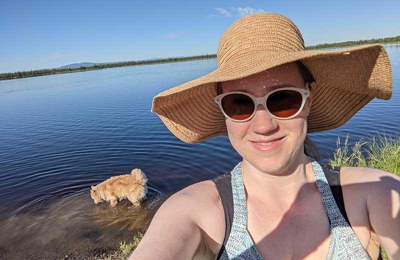 Woman with a hat and sunglasses stands by a lake with a dog. 