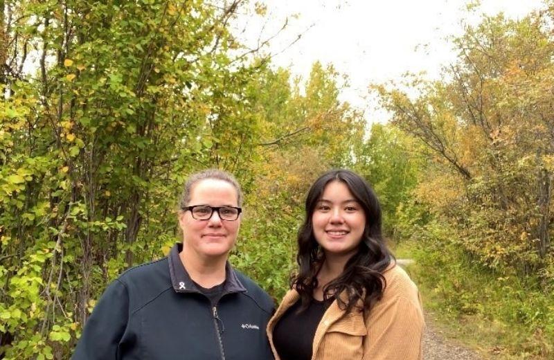 two women face the camera smiling there is a fall forest in the background