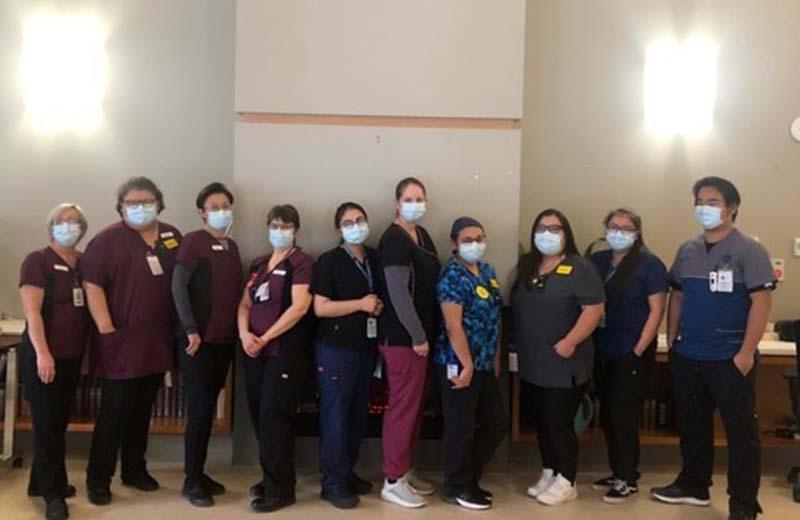 a group of health care professionals stand in a line to pose for a picture. they are wearing dark coloured scrubs and masks