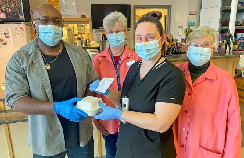 A group of people stand facing the camera wearing masks and medical scrubs. They are jointly holding a breakfast sandwich. 