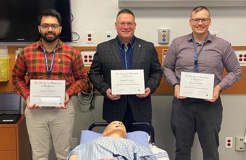 Three men stand in a row presenting their certificates over a medical dummy