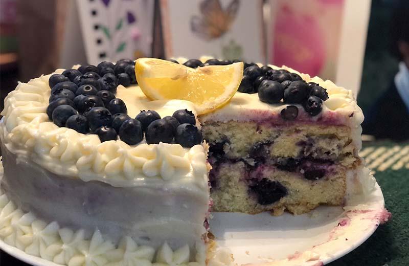 a white cake with white icing. there is a piece missing, you can see blueberries inside. On top of the cake is fresh blueberries and a lemon wedge.