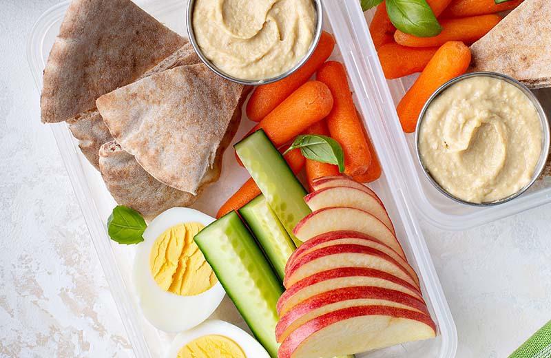 lunch container with sliced apple, hummus, pita bread, veggies and a boiled egg