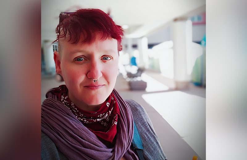 a person with short dyed-red hair smiles into the camera. They are wearing a cozy red scarf.
