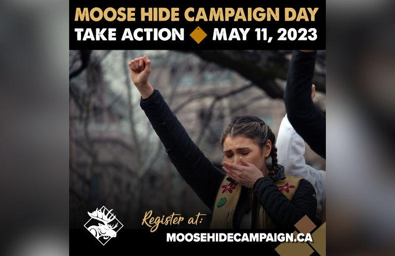 Moose hide campaign supporter standing with one hand over her mouth while the other is a raised fist in the air at a campaign event