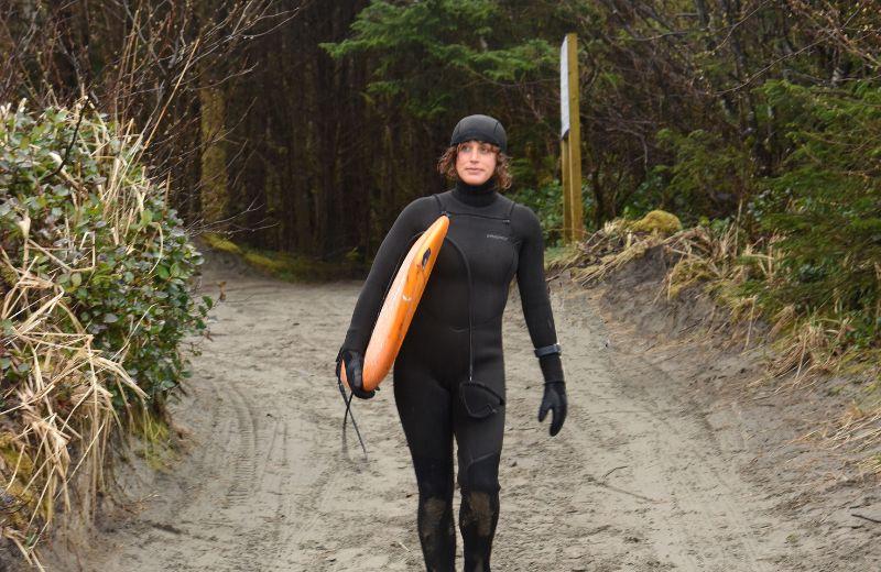 Dr. Jocelyn Black, head to toe in a black wet suit, walking down a beach pathway toward the water, holding her surf bard under her right arm.