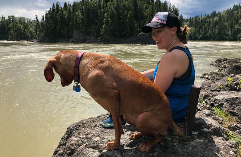 A woman sits with her dog on the shore of a river