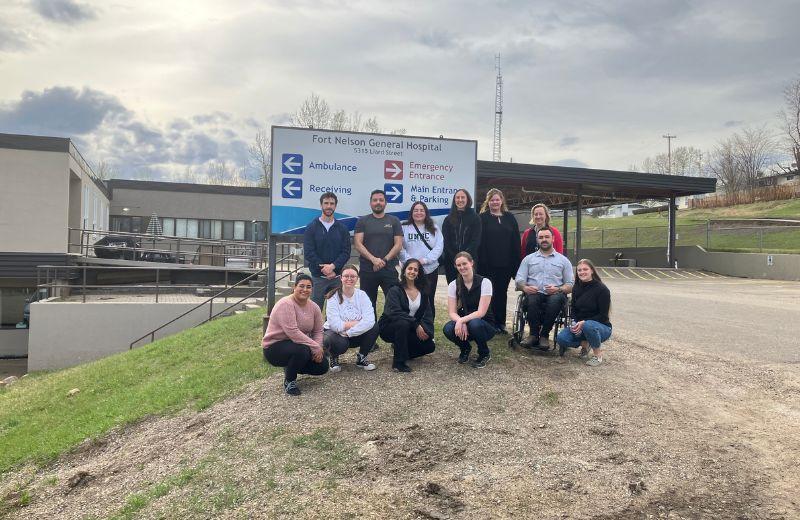 Students pose beside the entrance sign at the Fort Nelson Hospital