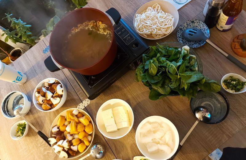 Several bowls containing noodles, tofu, greens, sauces and a large pot sitting on a table