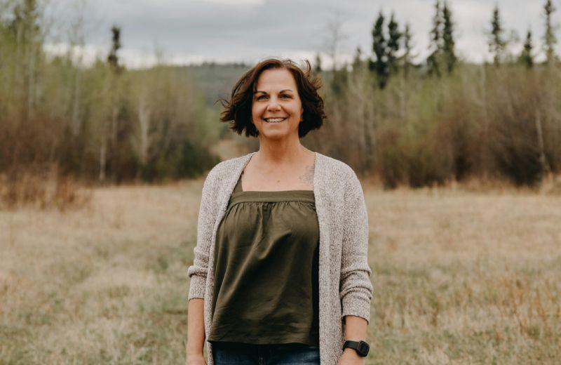 A woman in a green shirt with tan cardigan stands in a field in front of evergreen tress. 