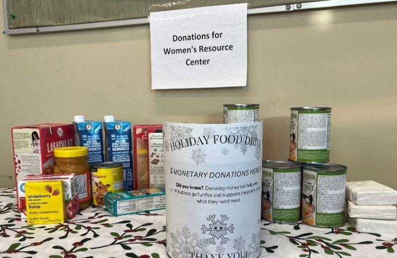 A stack of non-perishable food cans sits on a table covered with a holiday theme table cloth