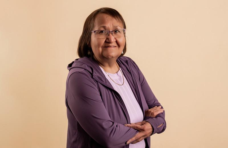 Indigenous woman stands with arms crossed across her chest and welcoming smile.