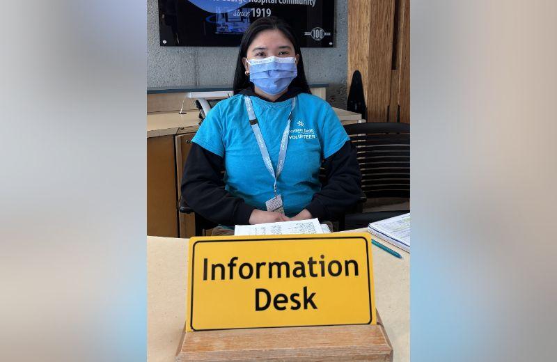 Diane sitting behind a yellow sign that reads "Information Desk"