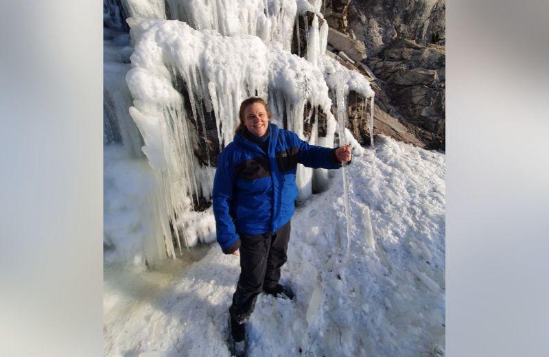 Penelope standing under a frozen waterfall wearing blue ice climbing gear