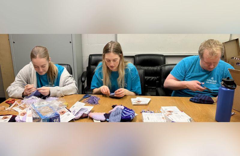 Volunteers knitting purple hats with supplies scattered across the table.