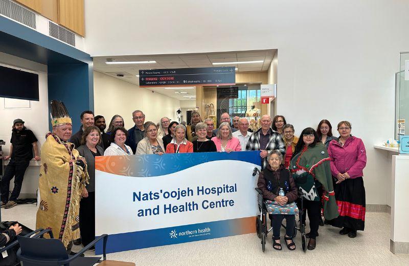 A large group of people stand holding a sign noting the name of the new hospital. 