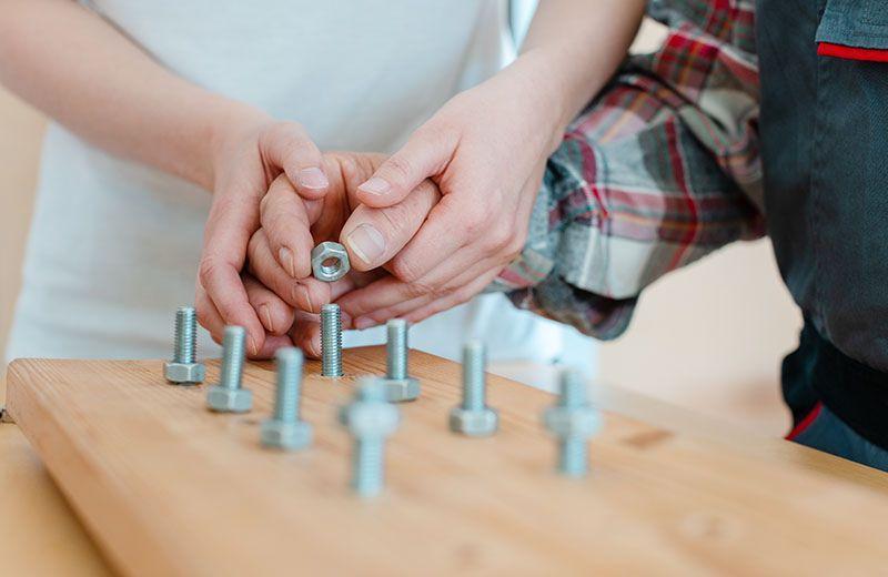 Two hands over a woodedn board attempting to screw nuts onto protruding bolts.
