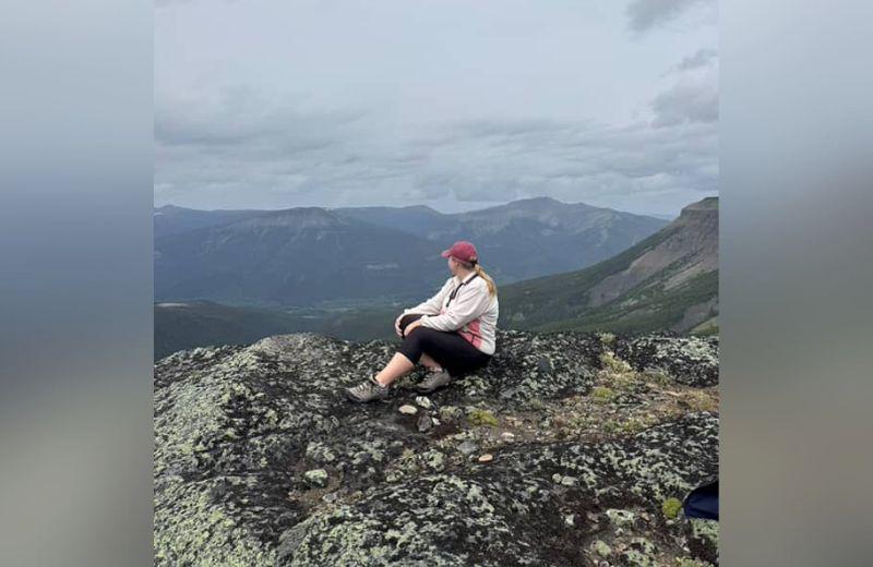 Amanda sits atop a mountain, viewing forests and mountains in the background. 