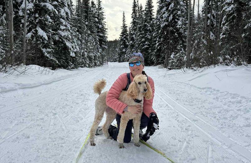 Haylee holding her dog on one knee in the snow, surrounded by trees