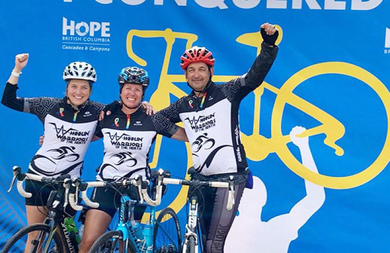Haylee, her mom, and dad celebrate finishing the 2019 Ride to Conquer Cancer in their bike outfits with helmets on, and arms raised. 