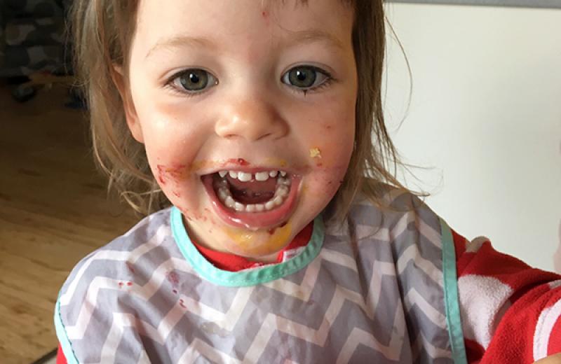 An adorable child, with food all over it's face, smiles into the camera and holds a peanut butter and jam sandwich. 