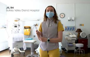 A picture of Jill - she is wearing yellow scrubs with a grey fleece vest on top. She has long light brown hair and is wearing a blue surgical mask.